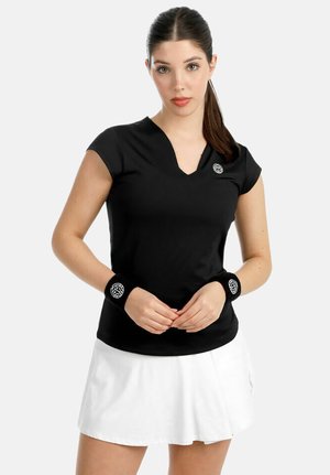 Frau trägt ein schwarzes T-Shirt mit V-Ausschnitt und kurzen Ärmeln, schwarze Handgelenkbänder mit Logos und weißen Rock, steht vor einfachem Hintergrund.