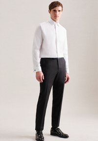 Camicia bianca con colletto alla francese, abbinata a pantaloni neri su misura e scarpe nere lucide. Tessuto liscio e strutturato.