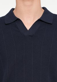 Laivastonsininen lyhythihainen poolopaita, jossa on teksturoitu neulontamalli, V-aukko ja tasainen kaulus. Kangas on resorikuosinen ja siinä on hienovaraisia pystysuoria viivoja.