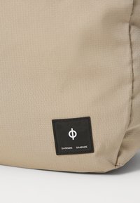 Beige stoffveske med et teksturert rutemønster. Har en svart etikett med hvit merkevaretekst og logo. Rund form, myke kanter.