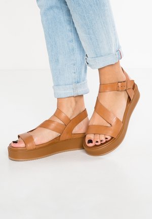 Pieds en jean bleu clair retroussé, portant des sandales à plateforme en cuir fauve avec des brides à la cheville et un vernis noir sur les ongles de pieds sur fond blanc.