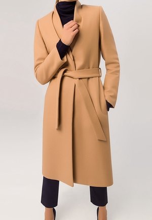 Long manteau beige ceinturé porté sur un col roulé noir et un pantalon noir court, une main ajustant le col et l'autre dans la poche.