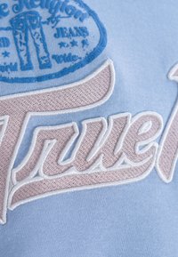 Le tissu bleu clair présente un logo brodé blanc avec des accents roses, mettant en valeur une écriture stylisée et un motif graphique rond au-dessus.