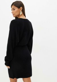 Abito nero lavorato a maglia con maniche lunghe e fluttuanti e una silhouette aderente. Presenta una texture liscia e un collo rotondo. Orecchini dichiarativi in oro.