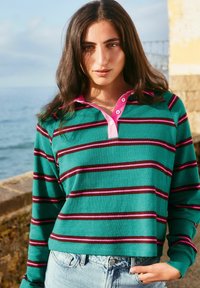 Green striped long-sleeve polo shirt with pink collar and dark maroon stripes, paired with light blue high-waisted denim jeans.