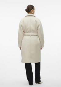 Crème kleurige wolmixjas met een ceintuur in de taille, een gesneden kraag en een gestructureerd oppervlak, gedragen over zwarte broek en crème hakken.