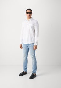 Weißes Langarmhemd mit einer Brusttasche, hellblaue verwaschene Jeans und schwarze Slipper. Person trägt dunkle Sonnenbrille.