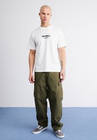 Camiseta blanca de algodón con el logo negro de "Dickies". Pantalones cargo verde oliva con bolsillos laterales. Zapatillas grises con suelas blancas. Diseño minimalista.