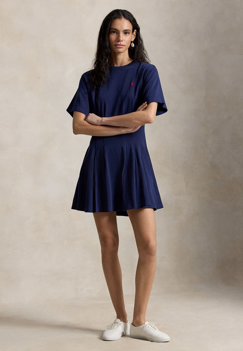 Junge Frau mit langen lockigen Haaren steht mit verschränkten Armen da und trägt ein marineblaues Kleid mit kurzen Ärmeln und weiße Turnschuhe vor einem neutralen Hintergrund.