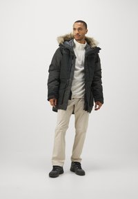 Sort isoleret parka med hætte med faux pelskant, frontlynlås og trykknapper. Bæres over en hvid fleece-top og beige bukser.