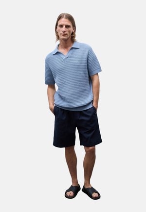 Homme debout les mains dans les poches portant un polo en maille bleu clair à manches courtes, un short foncé et des sandales noires sur un fond uni.