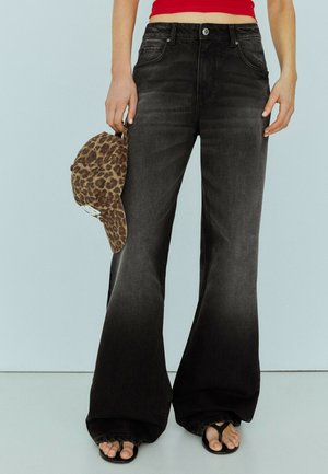 Person iført falmede sorte vidde jeans, sorte thongsandaler, rød cropped top, der holder leopardmønstret baseballkasket i venstre hånd.