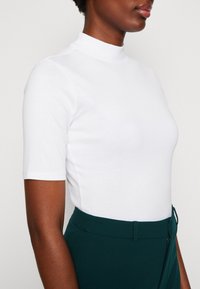 Hvid mock neck kortærmet top, tætsiddende design, glat tekstur. Sammen med højtaljede mørkegrønne bukser med en synlig lynlås.