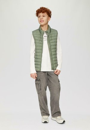 Grüne Pufferveste über einem weißen Langarmshirt, kombiniert mit grauen Cargo-Hosen und mehrfarbigen Turnschuhen. Weicher Stoff, lässiges Design.