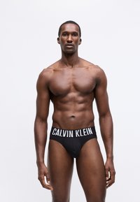 Svalnatý muž stojí vzpriamene, oblečený v čiernych slipoch Calvin Klein na jednoduchom bielom pozadí.