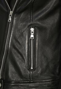 Gros plan d'une veste en cuir noire montrant deux fermetures éclair métalliques argentées, une fermeture éclair verticale de poche et une fermeture éclair frontale.