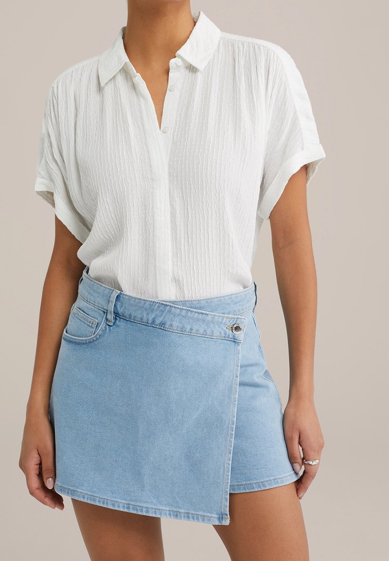 White textured blouse with a collar and roll-up sleeves, paired with a light blue denim wrap skirt featuring a side pocket and metal fastener.