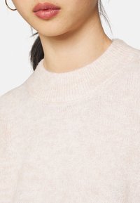 Pull rose clair avec un col montant côtelé et une texture en maille douce, mettant en valeur des fibres délicates et une coupe décontractée.