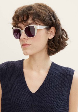 Lunettes de soleil avec une forme œil de chat, monture blanche et verres dégradés violet foncé. La tenue de la personne est un haut sans manches en tissu texturé bleu marine.