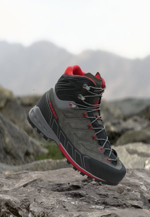 Szaro-czarny but turystyczny z czerwonymi akcentami i sznurowadłami, spoczywający na skalistym terenie z rozmytym górskim tłem.