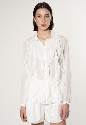 Weiße, transparente Bluse mit Rüschendetails entlang der Vorderseite und an den Ärmeln, mit einem Kragen und einer Knopfverschluss. Kombiniert mit weißen Shorts.