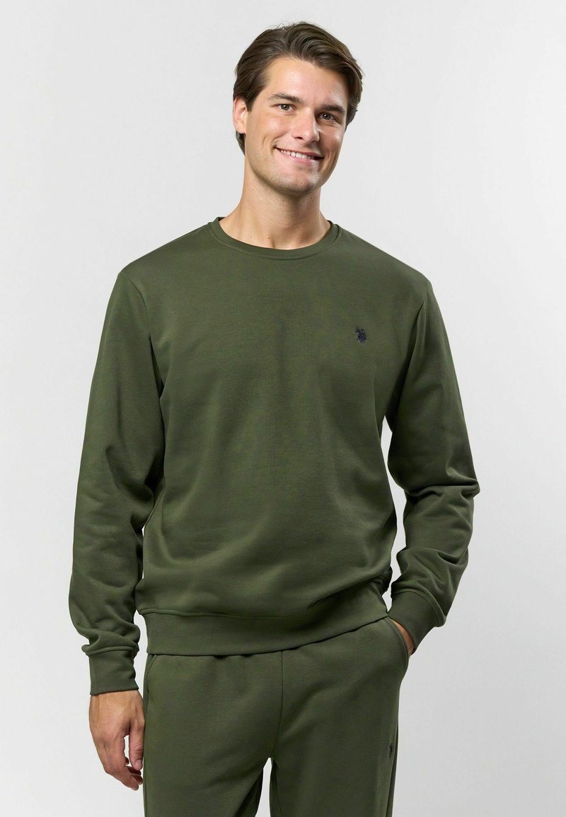 Olīvu zaļš džemperis, kas izgatavots no mīkstas auduma, ar apaļu kakla izgriezumu un garām piedurknēm, ar mazu izšūtu logo uz krūšu daļas.