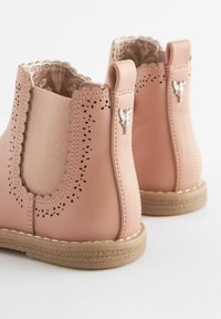 Botas de niño pequeñas en color rosa con bordes en forma de concha, detalles perforados, suelas beige y charms en forma de hada plateados en las lengüetas del talón.