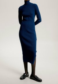 Persoon draagt een aansluitende navyblauwe geribbelde coltrui midi-jurk, zwarte doorschijnende panty's en zwarte leren loafers, staand tegen een effen achtergrond.