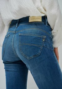 Jean en denim bleu avec une coupe ajustée, doté d'une patch en cuir avec logo, de poches arrière inclinées, de coutures contrastées et d'une texture lisse.