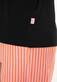 Černé tričko s malou růžovou nášivkou zobrazující ikonu zavřeného oka, doplněné oranžovými a růžovými kalhotami s vertikálními pruhy.