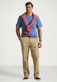 Blå stripete T-skjorte med rød strikkegenser bundet rundt halsen, beige bukser og marineblå sneakers. Brun belte med ur på håndleddet.