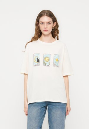 Mlada ženska z dolgimi valovitimi lasmi nosi belo majico z motivi v slogu tarotnih kart lune, sonca in zvezde, kombinirano z modrimi kavbojkami.