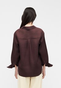 Femme aux longs cheveux foncés vue de dos portant une blouse ample marron foncé à manches longues et un pantalon beige clair sur un fond uni.