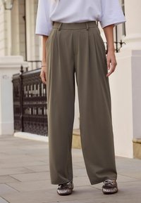 Leveälahkeiset housut oliivinvihreinä, joissa edessä laskokset ja korkeavyötäröinen malli. Yhdistetty kiiltäviin metallinhohtoisiin loafer-kenkiin. Sileä, kevyt kangas.