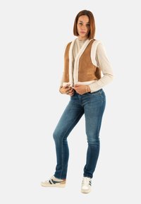 Brun mocka väst med vit shearling-fodring, bärs över en ljus tröja, matchad med blå denimjeans och beiga sneakers.