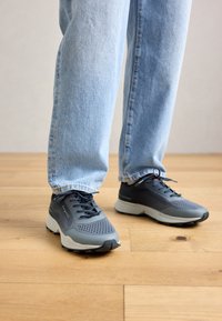 Pessoa vestindo jeans azul claro e sapatilhas Bugatti cinza-pretas, em pé sobre um chão de madeira clara.