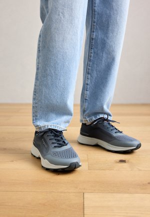 Osoba w jasnoniebieskich dżinsach i szaro-czarnych sneakersach Bugatti stoi na jasnej drewnianej podłodze.