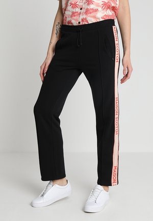Schwarze Hose mit pinken Seitenstreifen und dem Schriftzug "MAISON SCOTCH", getragen mit weißen Sneakern und einem pink gemusterten Hemd.