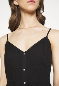 Vestido negro con escote en V, tirantes finos y tela texturizada que presenta pequeños círculos elevados. Cierre frontal con botones de acabado mate.