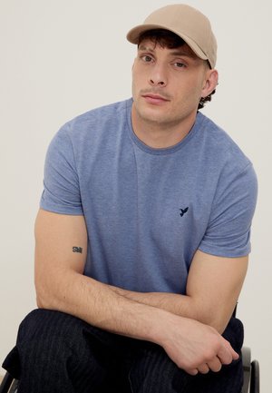 Άνδρας που φοράει μπεζ καπέλο και μπλε μπλούζα κάθεται με σταυρωμένα τα χέρια, δείχνοντας ένα μικρό τατουάζ στο δεξί του χέρι.