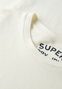 Cremerfarvet bomulds T-shirt med rund halsudskæring og blød tekstur; etiket med sort tekst delvist synlig på den indre hals.