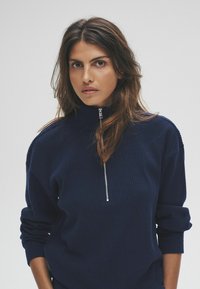 Femme aux longs cheveux bruns portant un pull côtelé bleu marine avec un col zippé, regardant directement l'objectif devant un fond uni.