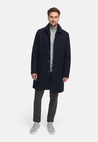 Marineblauer Wollmantel mit Kragen, vorderen Knöpfen und Seitentaschen, kombiniert mit einem grauen Rollkragenpullover, grauen Hosen und hellgrauen Sneakers.