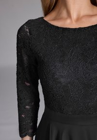 Top en dentelle noire à manches trois-quarts et col rond, avec des motifs floraux. Le bas est en tissu noir uni.