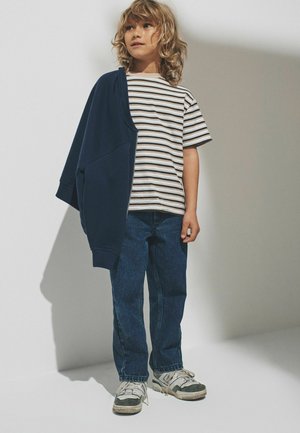 Kind mit lockigem Haar, trägt ein gestreiftes T-Shirt, blaue Jeans, weiße Turnschuhe und eine marineblaue Jacke, die lässig über eine Schulter gelegt ist, steht vor einem schlichten Hintergrund.