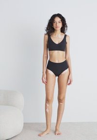 Black bikini set featuring a low-cut top and high-waisted bottoms. Curly hair, standing on a light carpet, with a minimalist background.