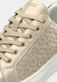 Beige sneaker with textured upper, logo detailing, and gold eyelets. Features flat beige laces and a white cushioned sole.