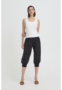 Vrouw staat met kort bruin haar, draagt een wit mouwloos topje, zwarte cropped broek en bruine open-toe hakken, tegen een effen achtergrond.