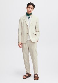 Ljusbeige linnekostym med en enkelknäppt kavaj och matchande byxor. Kombineras med en vit t-shirt och en grön mönstrad scarf. Bruna sandaler.