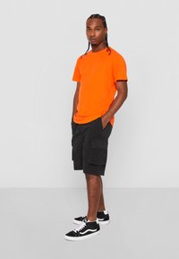 T-shirt orange vif à col rond, shorts cargo noirs avec deux grandes poches latérales, et baskets noires avec des accents blancs.